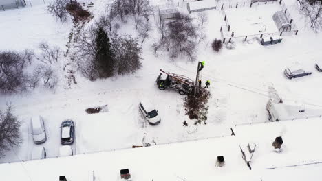 Maintenance-workers-replacing-electricity-pole-on-snowy-rural-village-by-the-apartment-building,-aerial-view