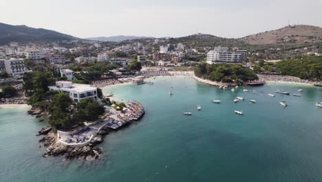 Aerial-view-of-picturesque-Ksamil-village-on-Albanian-Riviera's-Ionian-Sea-coastline,-showcasing-boats-in-the-tranquil-harbor