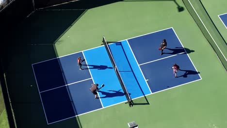 Pickleball-foursome-playing-on-sunny-day-hitting-ball-over-net,-aerial-descending-down-over-court