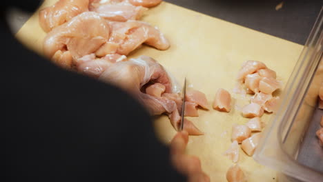 Chef-cuts-chicken-breast-into-bite-sized-pieces,-restaurant-kitchen-prep-of-dicing-chicken-on-cutting-board,-slow-motion-close-up-4K