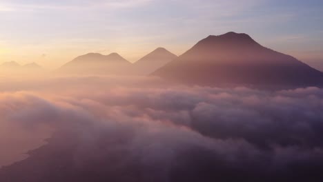 Epic-sunrise-drone-flight-at-cloud-level-over-San-Pedro-la-Laguna-around-Atitlan-Lake-in-Guatemala,-with-three-volcanos-visible-in-shot