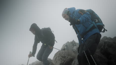 Two-hikers-deciding-what-direction-to-take,-climbing-down-from-the-rocks,-cloudy-and-foggy-weather-with-a-strong-wind