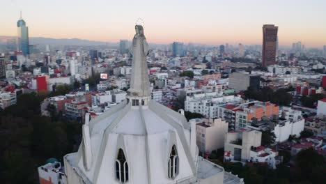 Breite-Drohnenaufnahmen-Der-Massiven-Jesus-Statue-Auf-Parroquia-Del-Purism-Corazon-De-Maria,-Mit-Mexiko-Stadt-Bei-Sonnenuntergang-Und-Einem-Park-Mit-Vielen-Bäumen-Im-Hintergrund