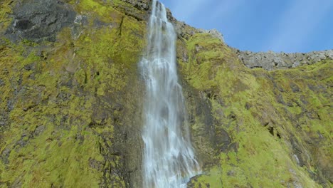 Looking-up-at-a-waterfall-flowing-over-green-moss-rock-on-a-bright-blue-day