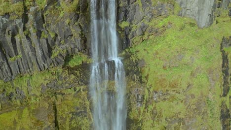 A-drone-follows-the-cascading-water-down-a-waterfall-over-a-green-and-yellow-cliff-face