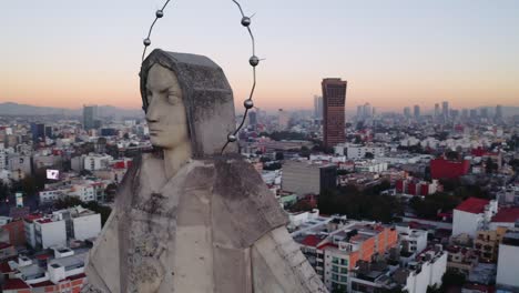 Dramatic-medium-drone-shot-of-massive-Jesus-status-towering-over-Mexico-City-at-sunset,-featuring-Parroquia-del-Purism-Corazon-de-Maria-and-buildings-in-the-back
