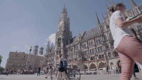 Low-angle-shot-of-people-walking-in-front-of-a-cathedral-in-Munich,-Germany-on-a-sunny-day,-enjoying-the-weather-in-a-documentary-style-handheld-shot,-everything-in-focus