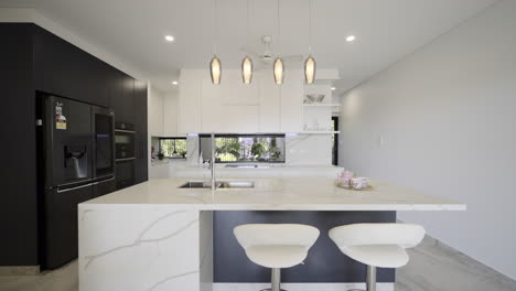 Modern-Luxurious-Contemporary-kitchen-white-marble-bench-top-drop-lighting-marble-tiled-floors-breakfast-bench-seating,-black-feature-wall-with-sleek-black-appliances-fridge-white-kitchen-storage