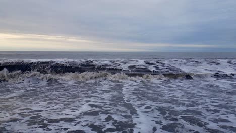 Grey-muddy-foaming-waves-crash-onto-a-black-sandy-beach