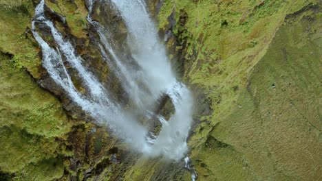 Establishing-shot-with-a-flyover-then-descending-down-a-large-waterfall-with-a-green-and-yellow-cliff-face-finishing-with-a-wide-pov