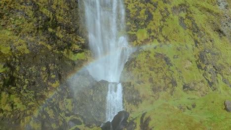 Wasser,-Das-Einen-Wasserfall-Mit-Einem-Regenbogen-Und-Grünem-Moos-Hinunterstürzt,-Das-Die-Felsige-Klippenwand-Bedeckt