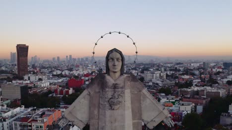 Medium-wide-backwards-flying-aerial-drone-shots-of-massive-Jesus-status-towering-over-Mexico-City-at-sunset,-featuring-Parroquia-del-Purism-Corazon-de-Maria-and-buildings-in-the-back