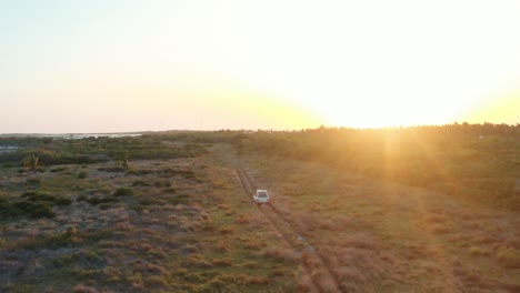 Very-wide-aerial-shot-of-all-terrain-SUV-with-a-rooftop-tent-riding-through-a-rough-field-towards-the-sunset