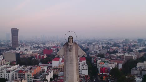 Close-up-drone-shots-of-massive-Jesus-status-towering-over-Mexico-City-at-sunset,-featuring-Parroquia-del-Purism-Corazon-de-Maria-and-buildings-in-the-back