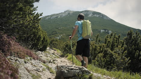 Camera-tracking-from-behind-a-hiker-with-a-hiking-poles-and-light-green-backpack-who-is-walking-up-on-a-rocky-path-towards-the-top-of-the-mountain-Snežnik