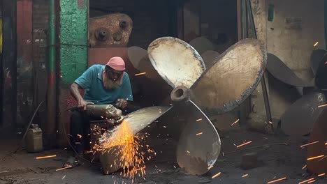 Old-man-worker-polishing-ship-propeller-by-angle-grinder-at-workshop-factory-in-Dhaka,-Bangladesh