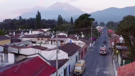 Colourful-chicken-bus-stops-to-pick-up-people-on-a-street-corner-in-Antigua,-Guatemala,-as-the-sun-rises-with-Acatenango-and-Fuego-volcanos-visible-in-the-background-and-birds-flying-in-front-of-drone