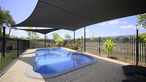 Large-luxurious-contemporary-outdoor-swimming-pool-in-ground,-clear-blue-water-black-fence-grey-concrete-decking-modern-home-green-grass-blue-skies-dark-shade-sails-pool-chairs