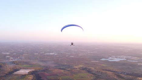 Powered-paraglider-soaring-high-above-Italian-countryside-during-bright-sunset,-aerial