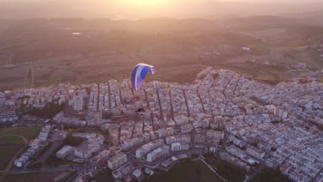 Paraglider-trike-freely-flying-above-Ginosa-in-Italy-during-bright-sunset,-aerial