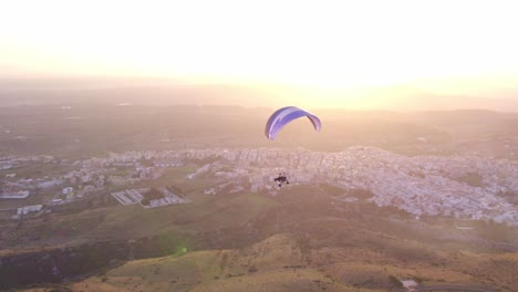 Powered-paraglider-soaring-in-sky-during-bright-sunset,-aerial