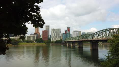 4K-Trucking-left-to-right-reveal-from-behind-a-tree-to-wide-shot-of-Hawthorne-Bridge-crossing-Willamette-River-toward-downtown-Portland,-Oregon-skyline-with-mostly-cloudy-sky