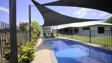 Large-luxurious-contemporary-outdoor-swimming-pool-in-ground,-clear-blue-water-black-fence-grey-concrete-decking-modern-home-green-grass-blue-skies-dark-shade-sails-outdoor-entertainment-area