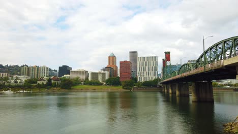 4K-Boom-up-reveal-from-behind-a-bush-to-Hawthorne-Bridge-crossing-Willamette-River-toward-downtown-Portland,-Oregon-skyline-with-mostly-cloudy-sky