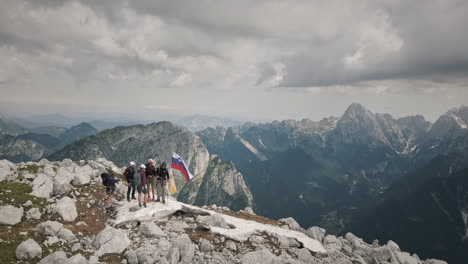 Drone-shot-from-top-of-mountain-Rombon,-a-group-of-hikers-with-a-slovenian-flag,-in-background-is-a-valley-and-mountains,-sky-is-covered-in-clouds