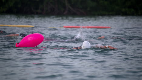 Female-athlete-swimming-in-a-triathlon-competition-wearing-a-cap-and-goggles-using-a-pink-buoy-for-support-looking-tired