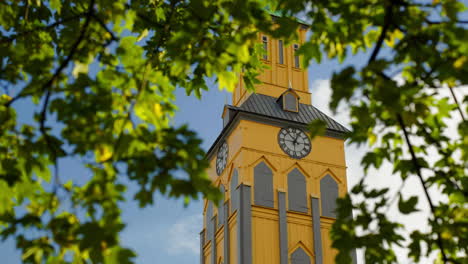 Domkirke,-Kathedralenfassade-Von-Tromso-Mit-Uhrturm-Hinter-Baumlaub-In-Tromso,-Norwegen-Low-Angle-Shot