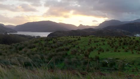 Ein-Starker-Wind-Bläst-Gras-Im-Vordergrund-Und-Wolken-über-Den-Himmel,-Während-Eine-Goldene-Sonne-Hinter-Bergen-Im-Hintergrund-In-Schottland-Untergeht