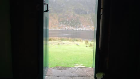 A-man-with-dreadlocks-and-wearing-hiking-gear-is-sitting-on-a-step-in-a-narrow-doorway-of-a-bothy-in-the-Highlands-of-Scotland-before-standing-up-and-walking-into-a-dark-room