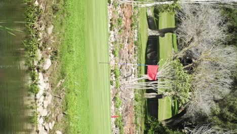 Vertical-Shot-Of-Green-Golf-Court-With-Red-Flag-In-Middle-Near-River