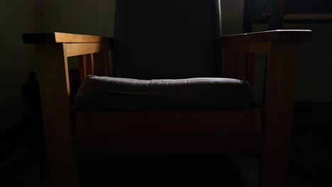 Eine-Langsame-Neigungsaufnahme-Zeigt-Einen-Alten-Gepolsterten-Sessel,-Der-In-Einem-Dunklen-Raum-Steht-Und-Von-Lichtstrahlen-Aus-Einem-Nahe-Gelegenen-Fenster-Beleuchtet-Wird