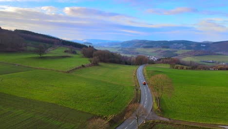 A-New-Day-in-Bruchhausen:-An-Aerial-View-of-an-orange-car-driving-a-land-road-with-the-Surrounding-green-Landscape-at-Sunrise