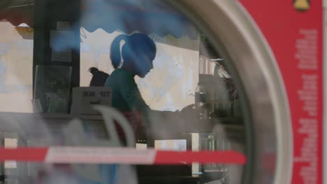 A-stationary-handheld-shot-of-a-reflection-from-the-glass-surface-of-a-front-load-washing-machine-shows-a-sitting-woman-browsing-her-phone-inside-the-laundry-shop-while-waiting-for-her-clothes