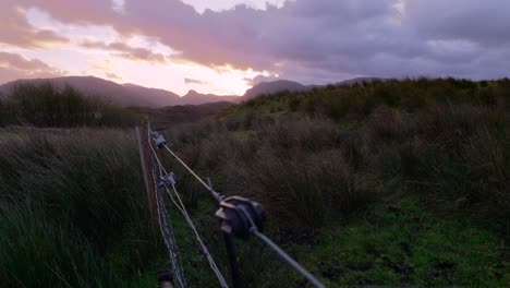 A-time-lapse-of-pink-and-yellow-clouds-flying-past-in-high-winds-at-sunset-with-a-livestock-fence-and-tufts-of-grass-sit-in-the-foreground-and-mountains-are-silhouetted-in-the-background