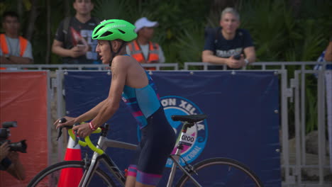 Latin-teenage-boy-competitor-getting-off-his-bike-barefooted-while-entering-the-transition-zone-at-a-triathlon-competition-wearing-a-green-helmet