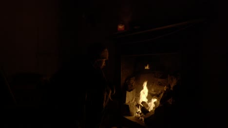A-man-sits-in-a-dark-room-warming-himself-by-a-log-fire-in-an-old-building