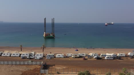 A-jackup-rig-or-a-self-elevating-unit-mobile-platform-in-an-offshore-area-in-front-of-Public-HaDatiyim-beach-Camping-site-on-Sunny-day,-Eilat-City,-Israel---aerial-dolly-right