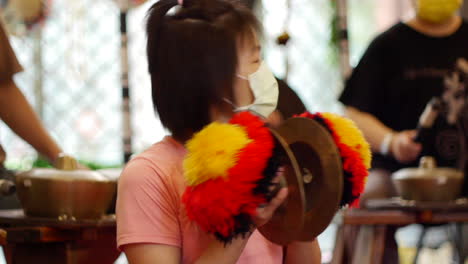 Asian-woman-wearing-face-mask-playing-decorated-cymbals-by-hand-during-performance,-filmed-as-medium-close-shot