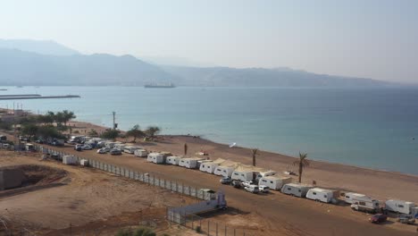 -HaDatiyim-beach-camping-site-with-many-caravan-cars-parked-at-the-coastline-on-a-sunny-day,-Eilat-City,-Israel---aerial-dolly-right,-Revealing-self-elevating-platform-for-shipment-in-shallow-waters