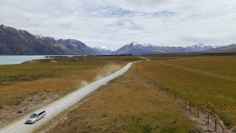 Silver-van-on-dusty,-picturesque-dirt-road-with-snow-capped-mountains-in-background