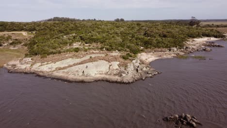 Two-people-walking-over-rocks-along-Black-Lagoon,-Punta-del-Diablo-in-Uruguay
