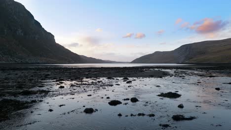 Water-slowly-flows-into-a-sea-loch-at-low-tide-in-the-highlands-of-Scotland-at-sunset,-surrounded-by-mountains