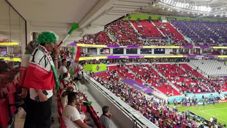 Ein-Paar,-Das-Die-Iranische-Fußballmannschaft-In-Doha,-Katar-Unterstützt,-Singen-Und-Schreien-Fröhlich-Mit-Weiß-grün-roten-Fahnendesign-kostümen-Und-Lockigem-Haardesign-Mit-Hijab-Im-Thumama-stadion-Der-Luxusgegend