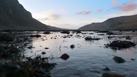 A-low,-sealevel-view-of-seaweed-and-water-slowly-flowing-into-a-sea-loch-at-low-tide-in-the-highlands-of-Scotland-at-sunset,-surrounded-by-mountains