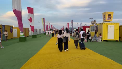 two-women-with-dark-black-hairs-white-shirt-and-black-trousers-are-walking-in-a-yellow-green-floor-in-the-fan-village-in-Doha-Qatar-designed-with-flags-and-suits-for-accommodation-in-a-colorful-place