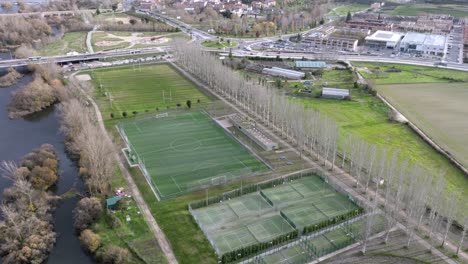 Volando-Sobre-El-Campo-Verde-De-Fútbol,-Tenis-Y-Canchas-De-Atletismo-Cerca-Del-Río-Azul-En-España